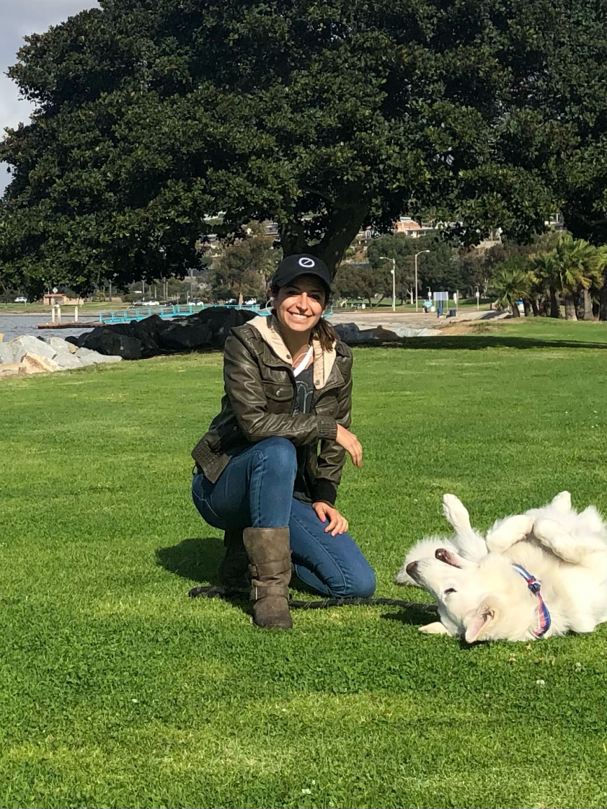 Mary, a Tender Hearts Dog Rescue teammate, kneeling on the grass in a park near the water with a large tree behind her while her white Husky is rolling in the grass.